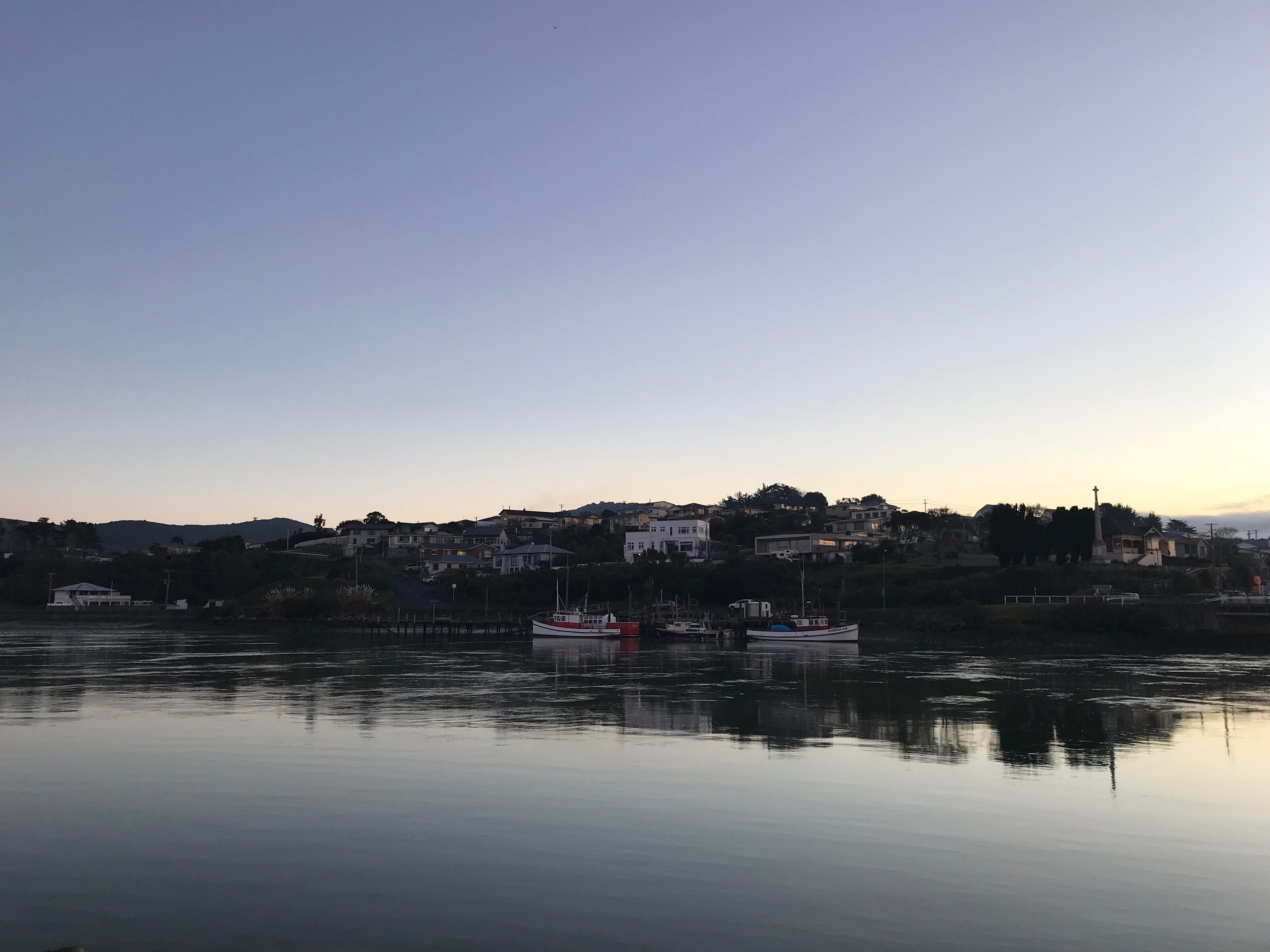 Fishing boats moored in Riverton Harbour at evening light, with calm reflections on the water and houses along the hillside.