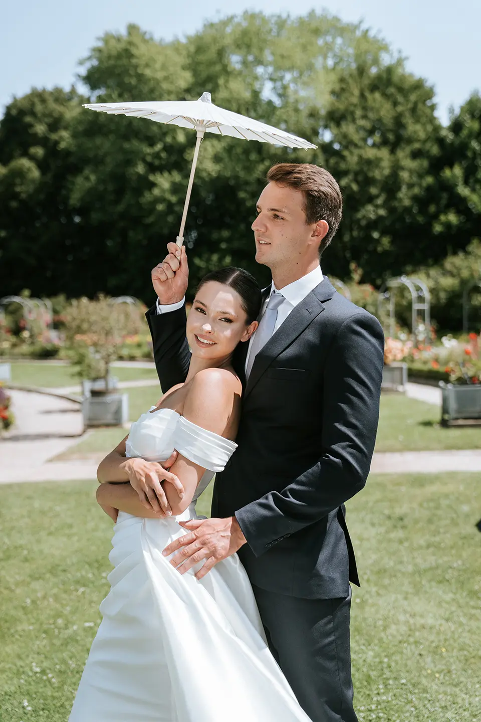 Eng umschlungen steht das Paar im sonnigen Garten unter einem Papierparasol, zarte Beete dahinter; fotograf für sommerhochzeit, natürliche hochzeitsfotografie und pärchenshooting hochzeit
