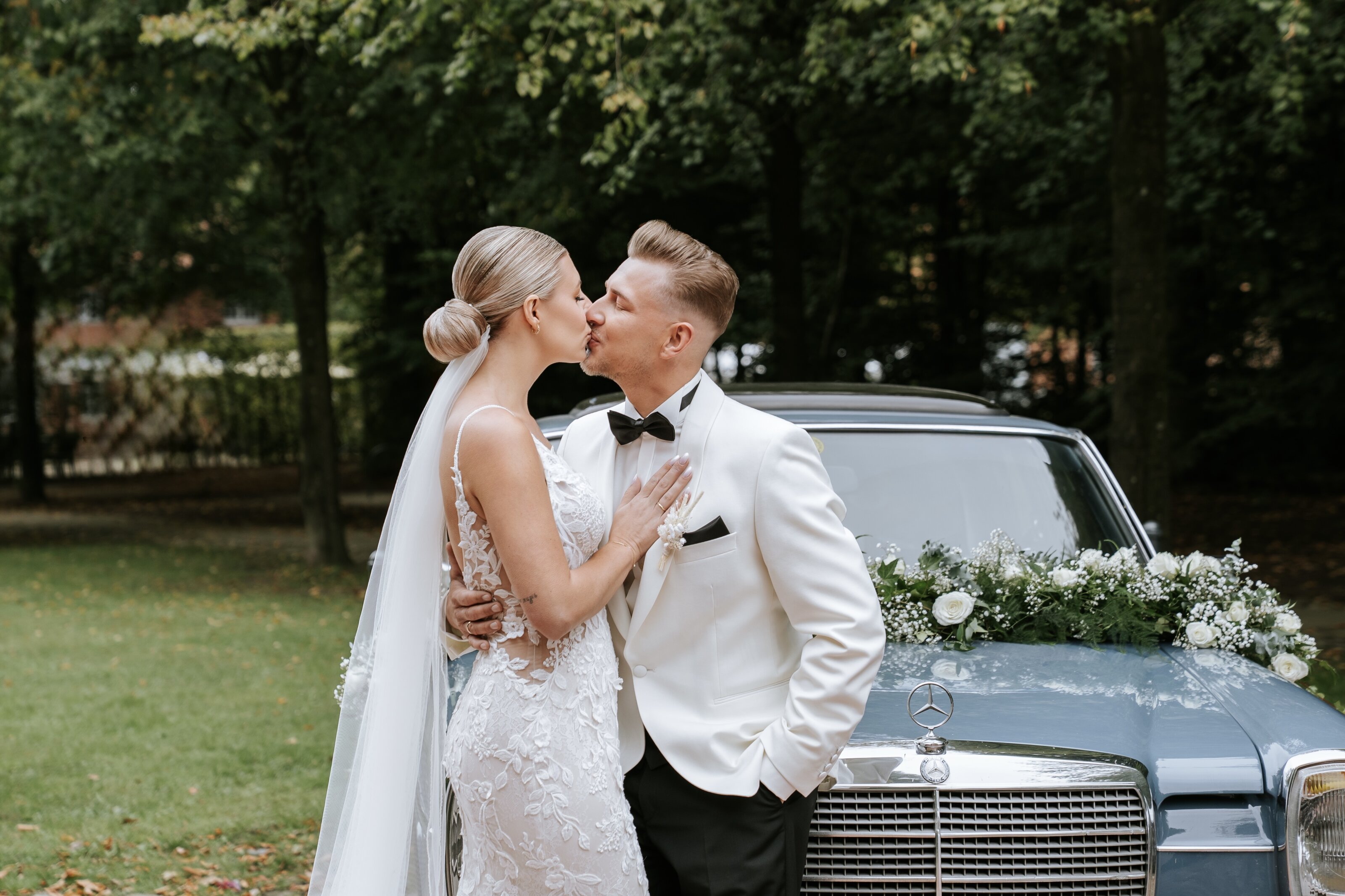 Das Paar spaziert über eine sonnige Wiese, sie im Spitzenkleid mit weißem Strauß, er im hellen Anzug. Natürliche Hochzeitsfotografie und hochzeitsbilder draußen mit dezentem Stil