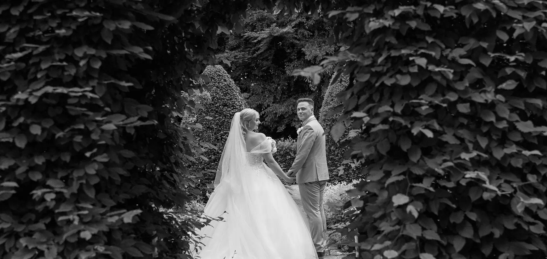 Brautpaar Hand in Hand im Garten, von dichtem Heckenbogen eingerahmt; eleganter Blick zurück in zeitloser Schwarzweiß, natürliche hochzeitsfotografie und hochzeitsbilder draußen