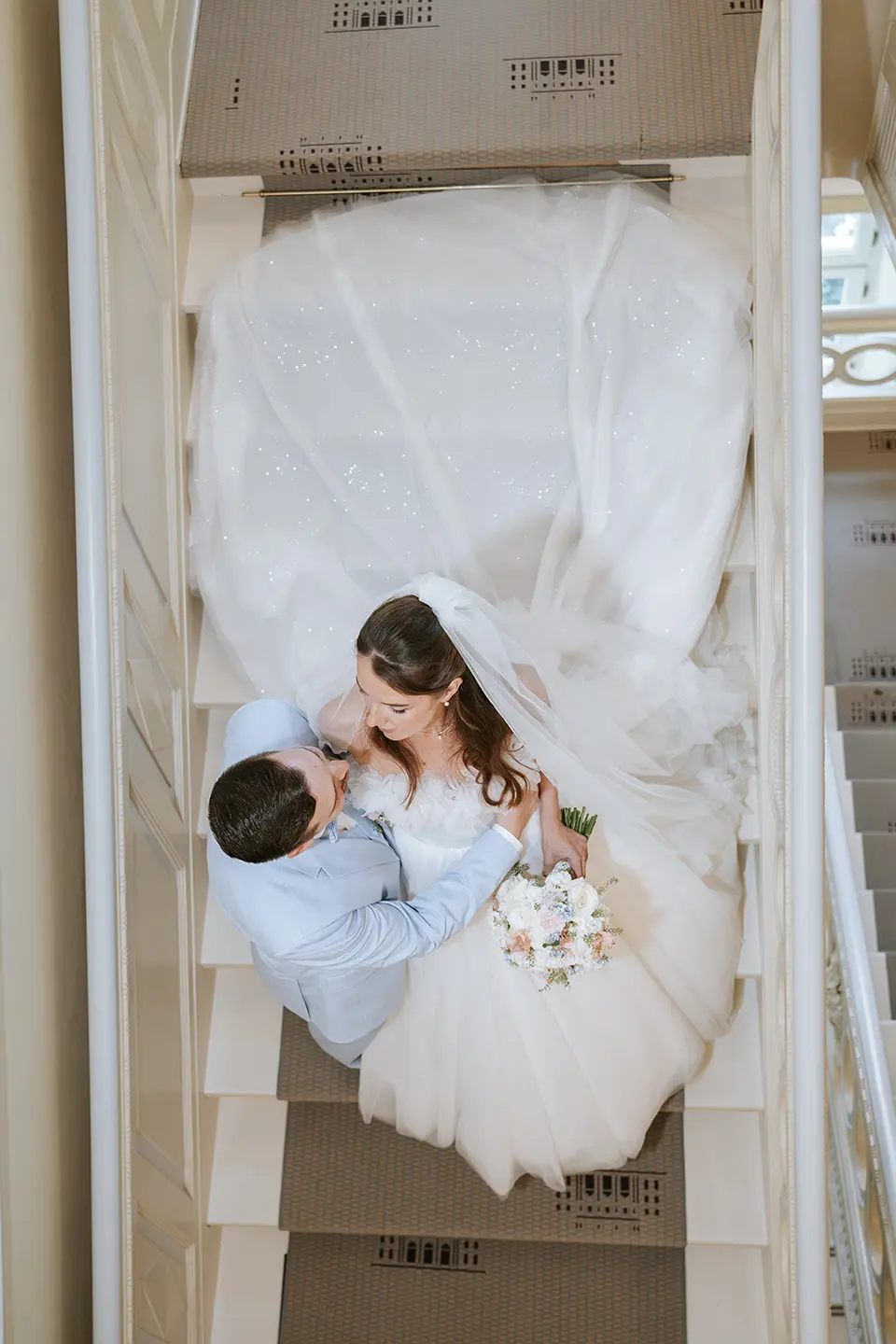 Aufsicht auf das Paar auf der Treppe, das Kleid fächert sich großzügig aus, der Strauß ruht in ihrer Hand; künstlerische hochzeitsfotos und moderne hochzeitsfotografie