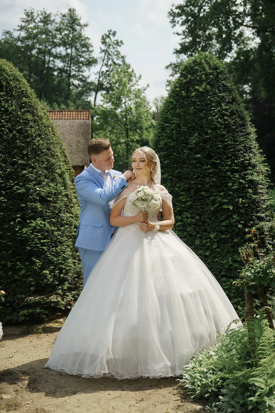 Bräutigam richtet der Braut zärtlich das Kleid, sie hält den Strauß zwischen hohen Hecken; warmer Gartenmoment für pärchenshooting hochzeit, natürliche hochzeitsfotografie und emotionale hochzeitsbilder