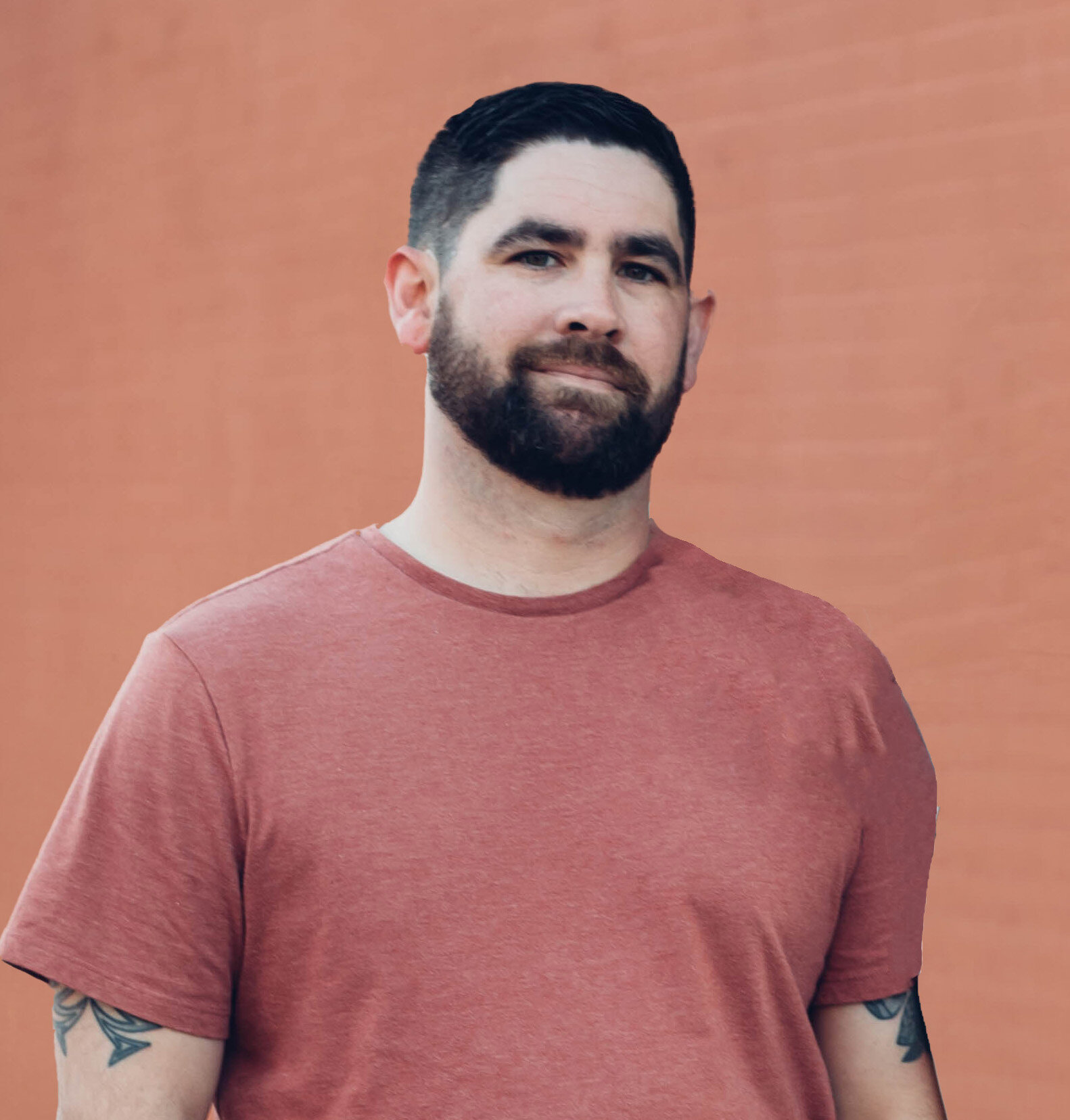 Man with short dark hair and beard wearing a pinkish-red t-shirt stands in front of a plain light red brick wall, embodying the creative spirit of a Showit designer specializing in brand and web design for small businesses.