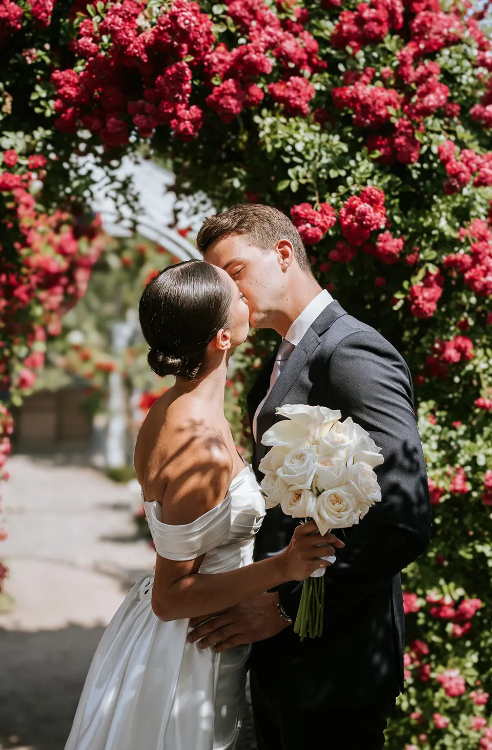 Zwischen dichten Rosen küsst sich das Paar, die Braut hält einen hellen Rosenstrauß; künstlerische hochzeitsfotos und moderne hochzeitsfotografie mit sanftem Gegenlicht