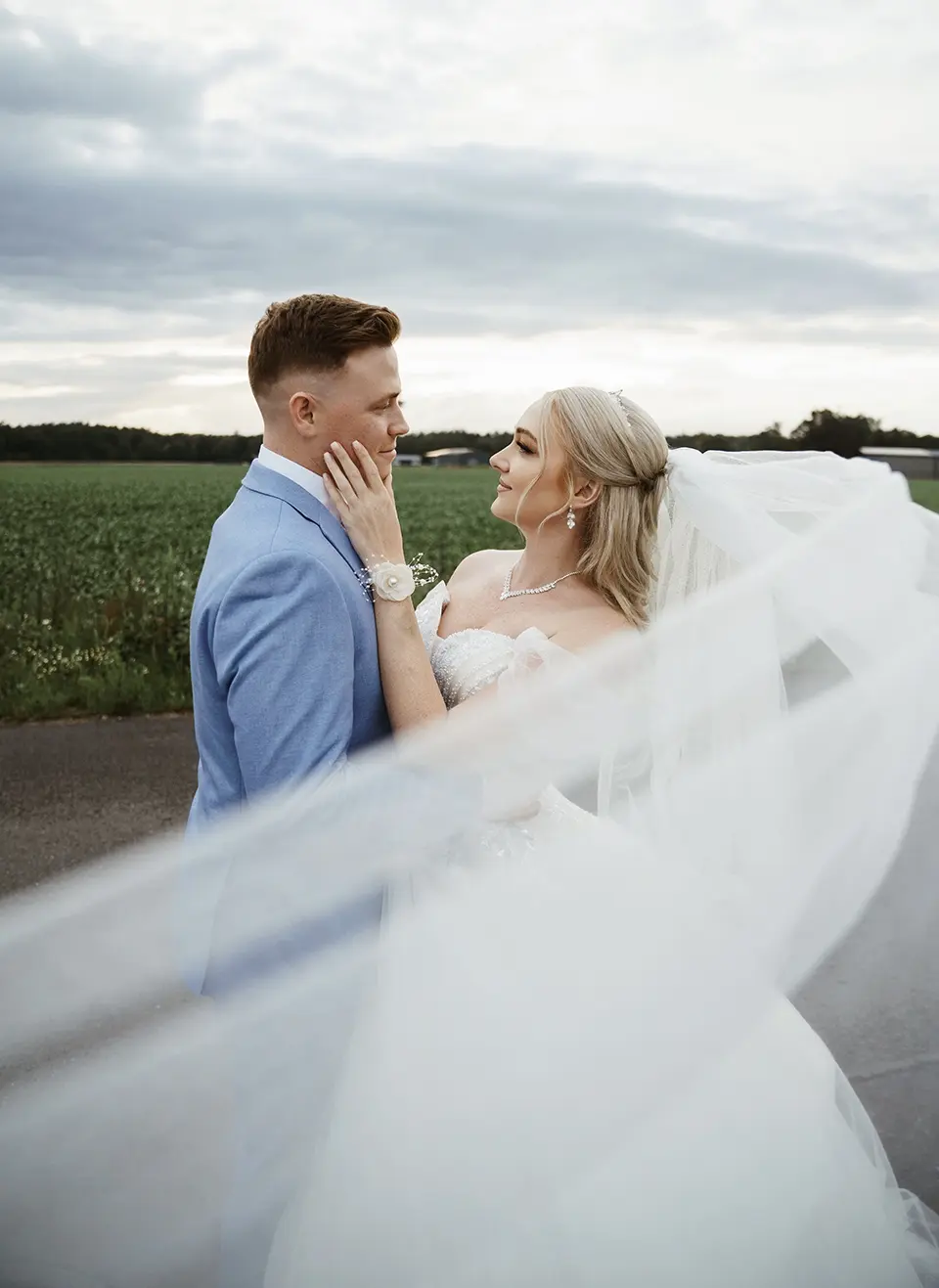 Das Brautpaar steht eng umarmt am Feldweg, der lange Schleier weht im Abendwind; sanfter Blickkontakt – pärchenshooting hochzeit und emotionale hochzeitsbilder, natürliche hochzeitsfotografie
