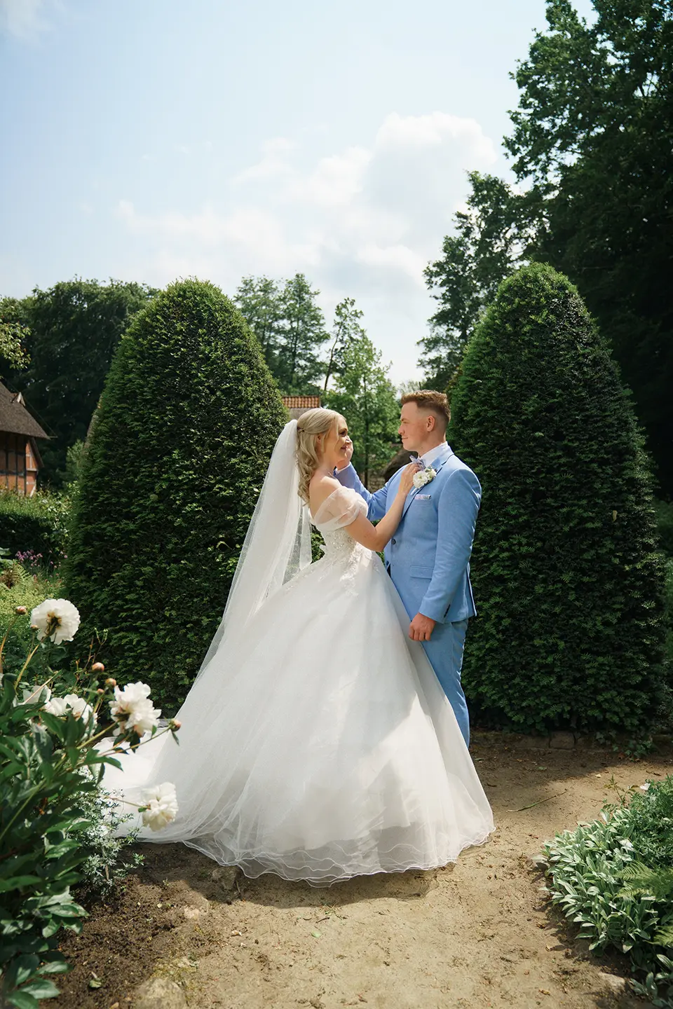 Braut im Prinzessinkleid und Bräutigam im hellblauen Anzug stehen zwischen hohen Buchsbaumkegeln. Hochzeitsbilder draußen und natürliche hochzeitsfotografie im Sommergarten