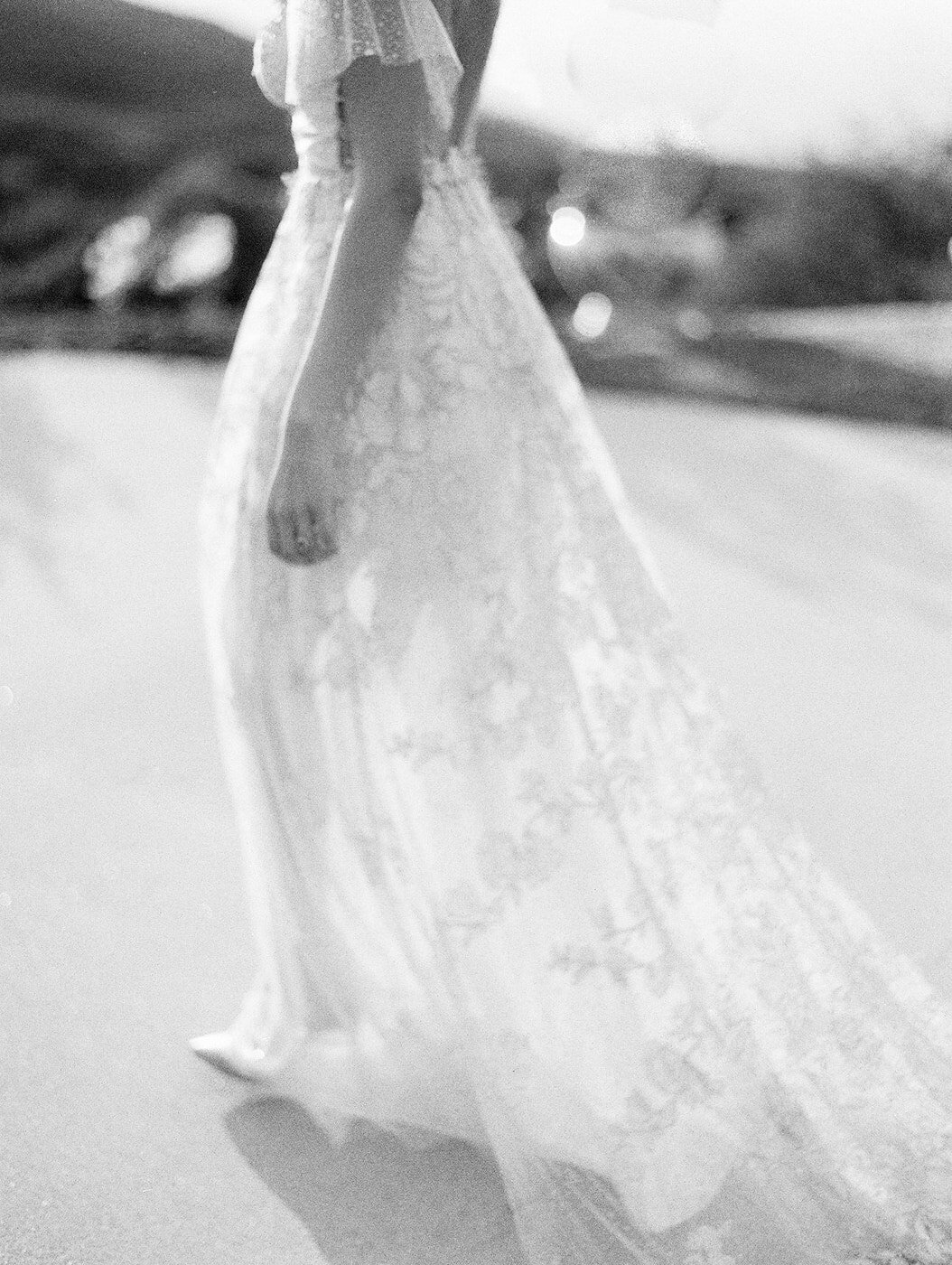 a grainy, slightly motion-blurred, black and white film photo of a bride wearing a bridal gown and heals. She is walking away so you can see the dress's intricate floral details and it was captured at Stone Mountain Estate in Malibu