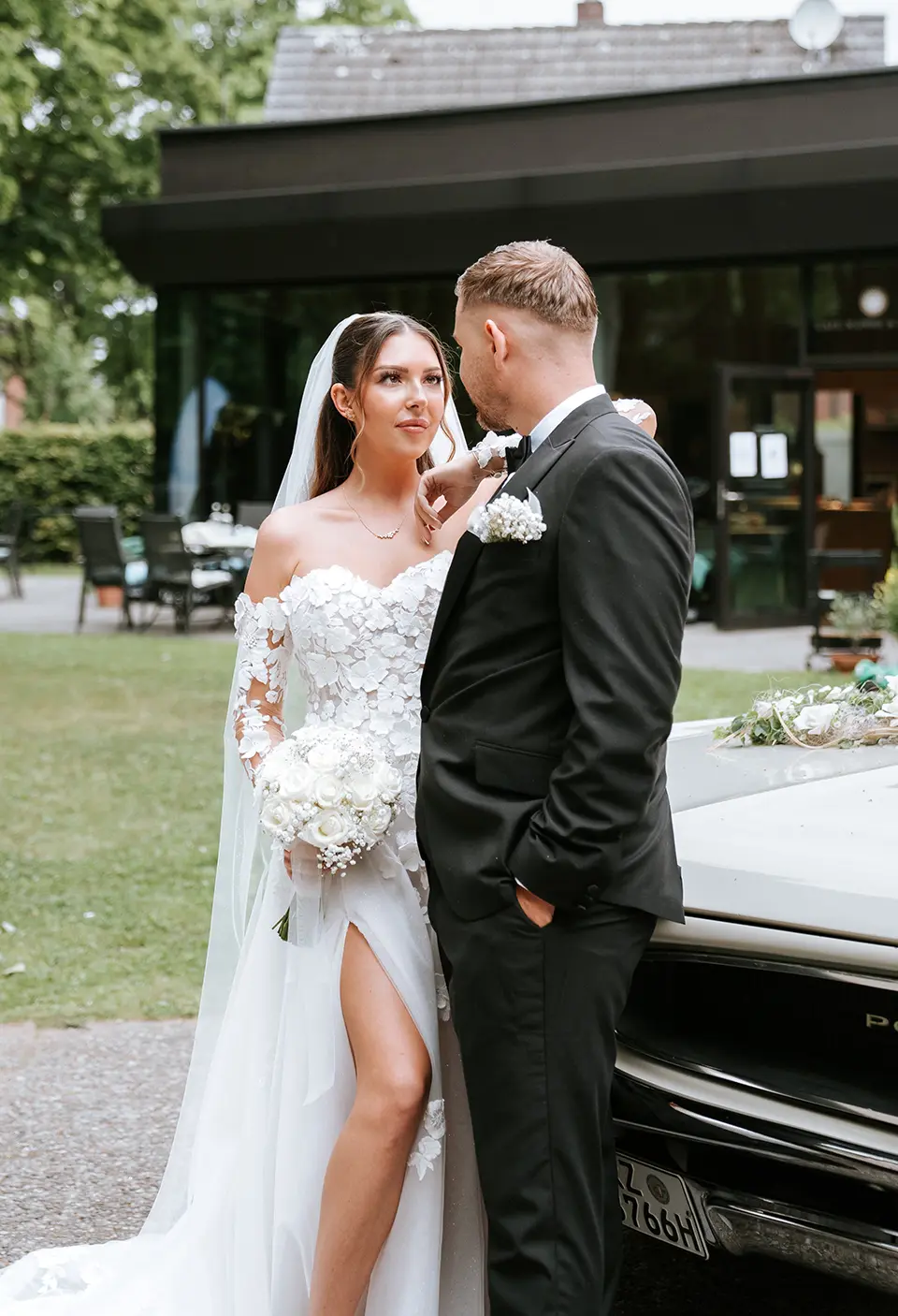 Neben dem Oldtimer blickt das Paar sich tief in die Augen, die Braut hält einen weißen Strauß; hochzeitsfotografie mit stil und erfahrener fotograf für hochzeit
