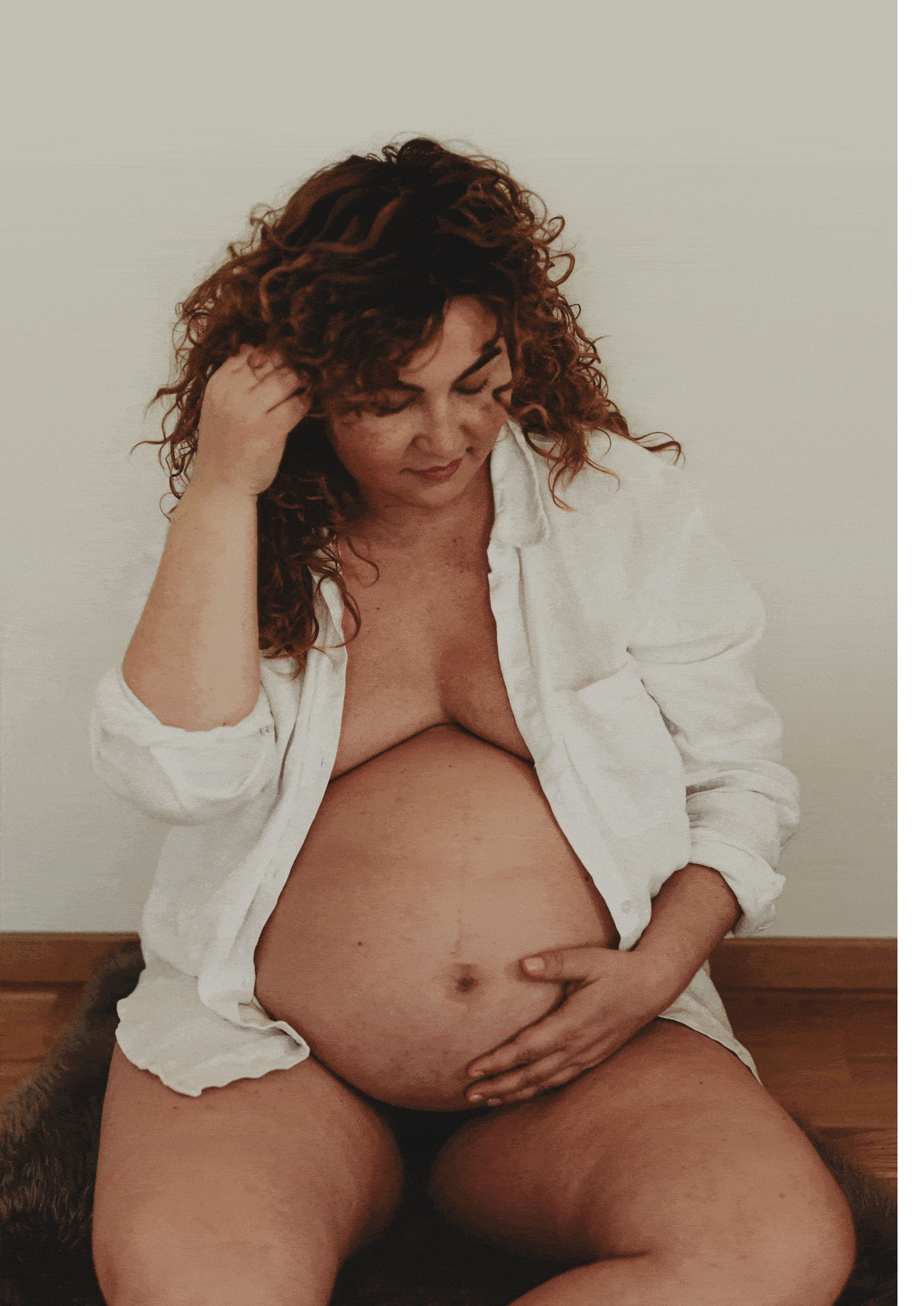 Gif-fil på en gravid kvinna sittandes på golvet iklädd vit skjorta hållandes en hand på sin gravida mage under en gravidfotografering i Stockholm