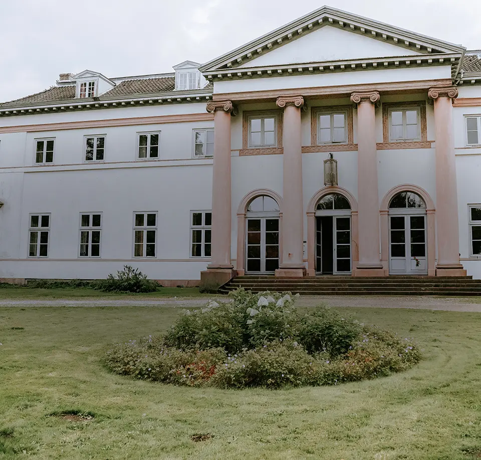 Weißes Herrenhaus mit rosafarbenen Säulen, symmetrischen Fenstern und Rasenrondell – ein einladender Ort für eine Schloss-Hochzeit, perfekt eingefangen in Hochzeitsfotografie mit Stil