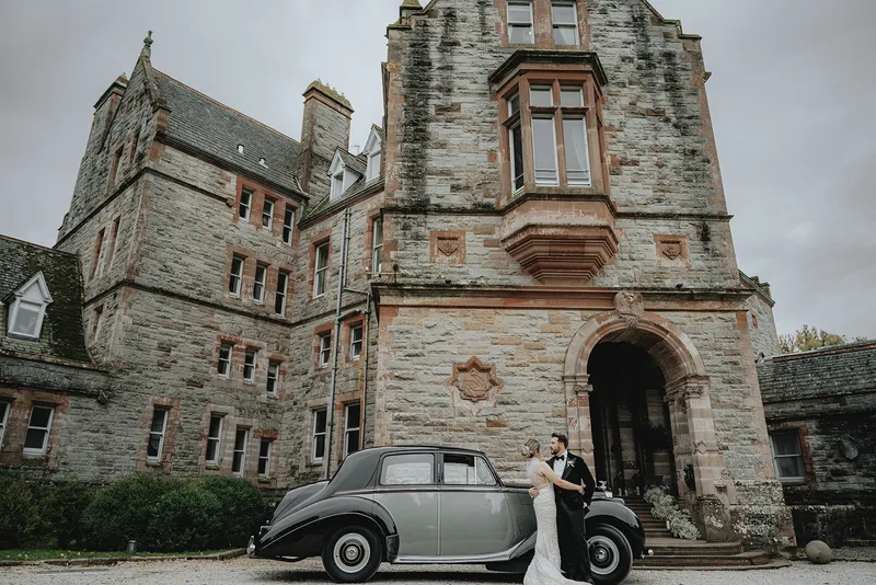 Couple beside a vintage car outside the stone entrance of Castle Leslie Estate in Ireland.